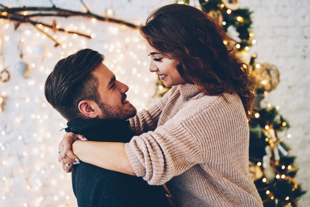 A couple embraces while smiling. Christmas decorations are seen behind them.