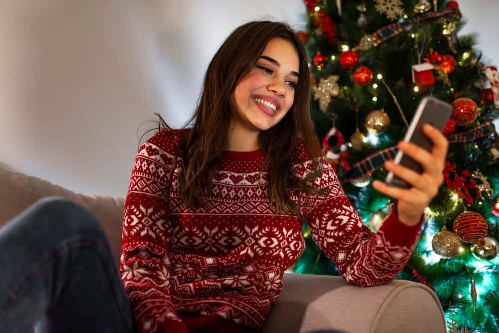 A woman smiles at her phone reading something while in a Christmas sweater.