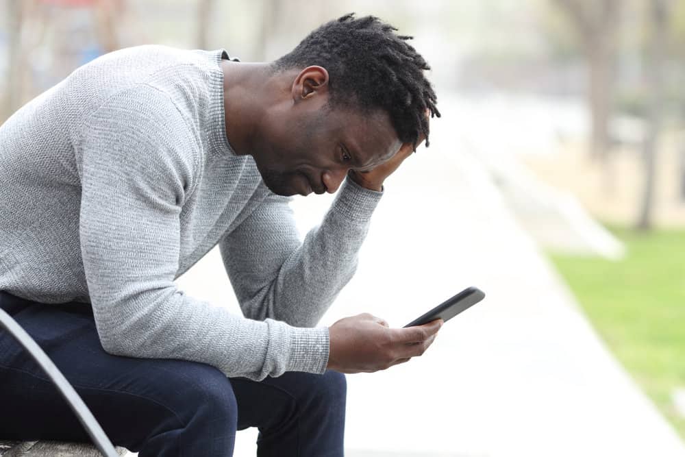 A man with a distraught face holds his head while reading a message on his phone outdoors.