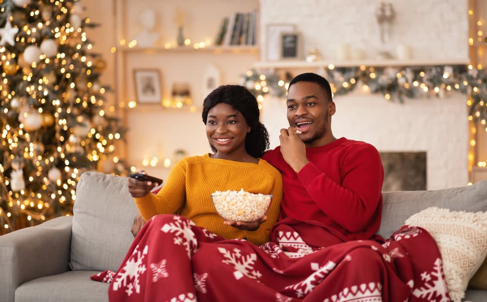 A couple smiles on the couch sharing a blanket while holding popcorn.
