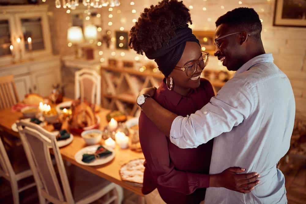 A couple embraces with a Thanksgiving table set behind them as they smile.