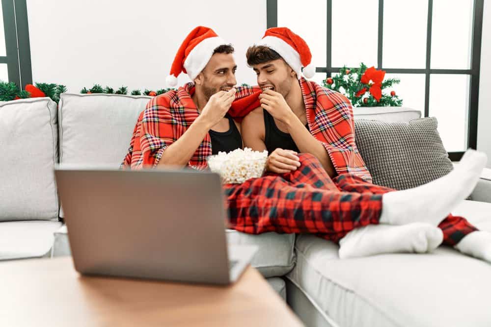 A couple sits on a couch while eating popcorn. They watch something on a laptop which is in front of them.