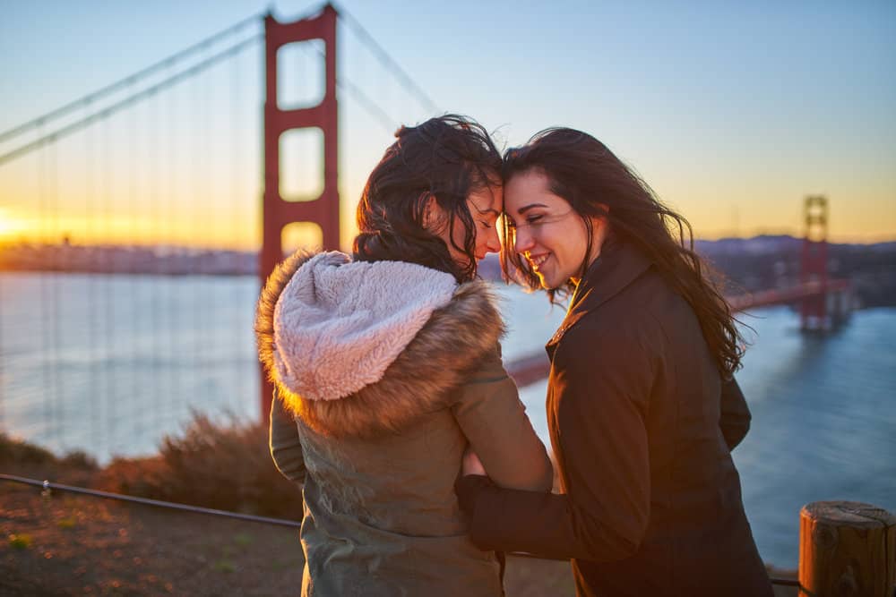 Two women stand forehead to forehead in San Francisco with a big red bridge behind them.