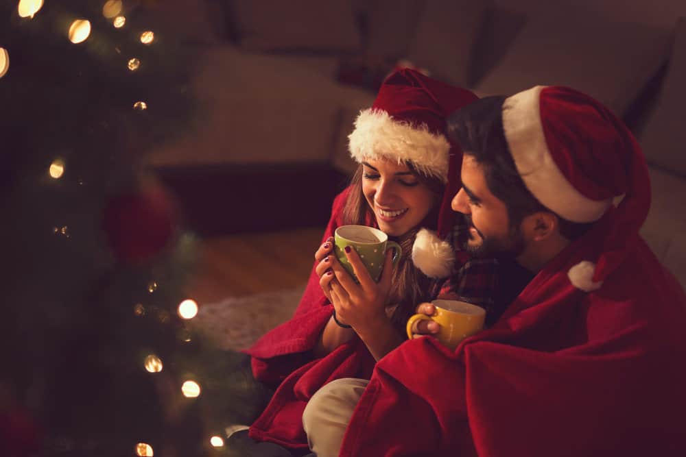 A couple snuggles in Christmas hats by the tree with mugs in their hands.