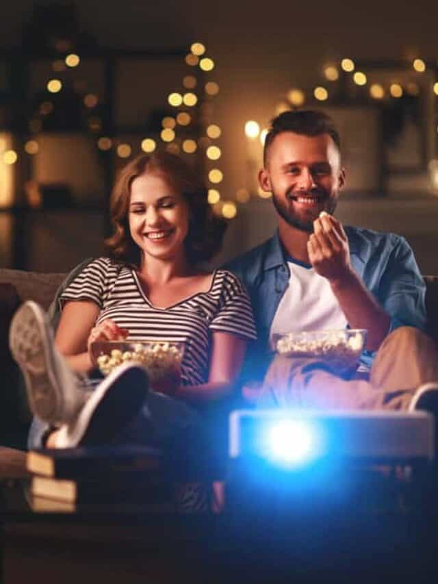 A couple sitting together eating from small bowls of popcorn with a projector placed on the table in front of them.