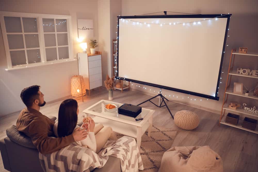 A couple sitting on a couch in their home in front of a small projector screen with string lights hanging around it. 