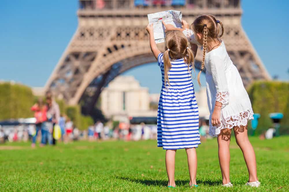 Two children holding a map up and pointing at it while they stand in a park at the foot of the Eiffel Tower. 
