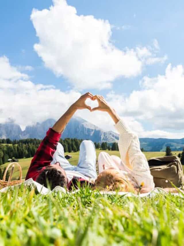 A couple lays on their backs in a field while making a heart with their hands, planning a romantic getaway together