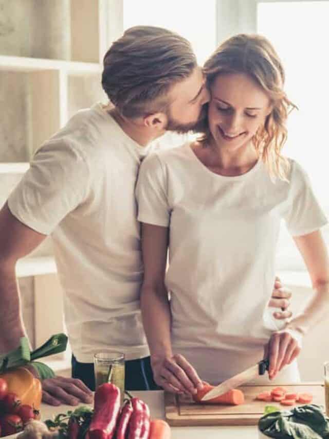 A man kisses his spouse on the cheek as she chops vegetables in a sunny kitchen while discussing how to manage expectations in marriage