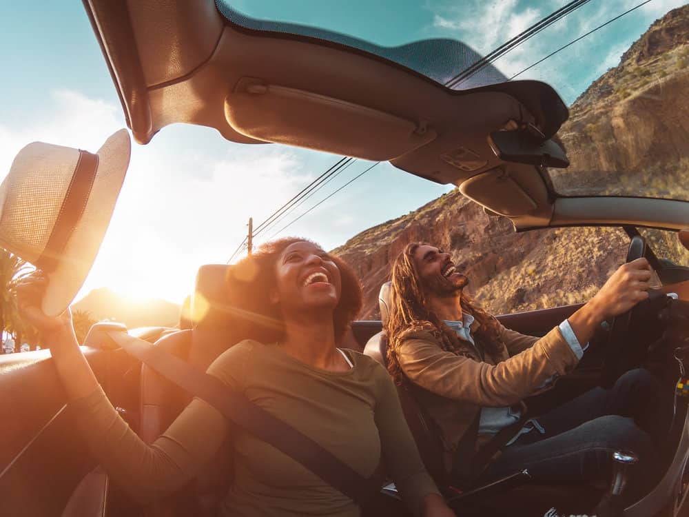 A couple is smiling while in a convertible driving through the streets.