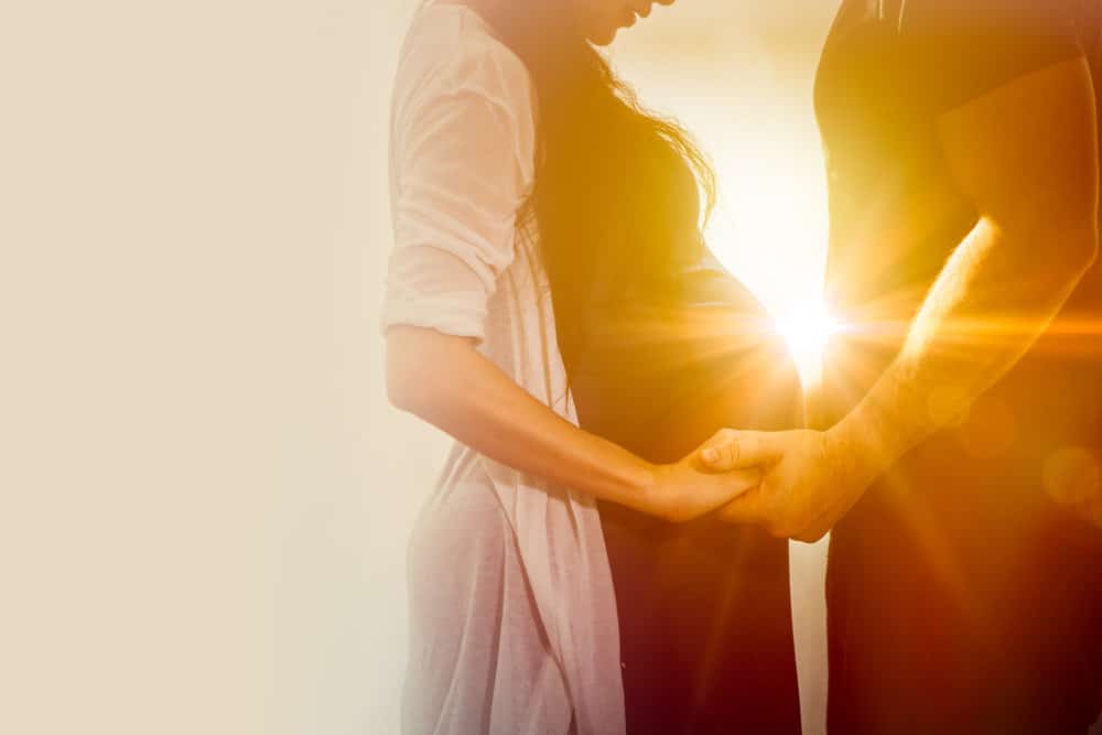 A pregnant couple holding hands as the sun shines in between them.