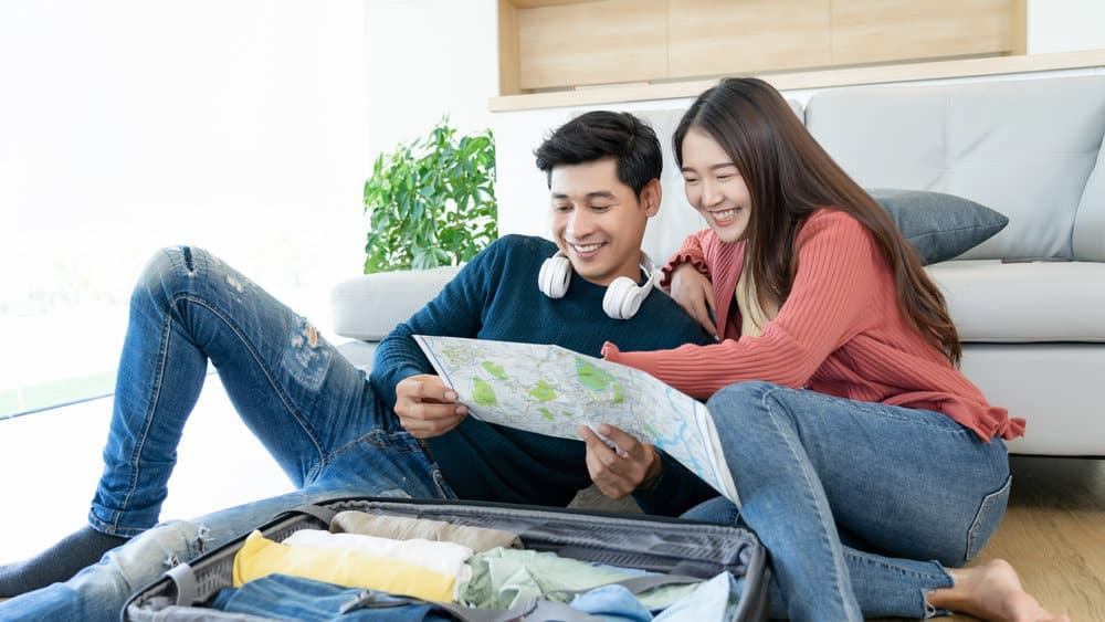 A couple smiles while looking at a map next to a suitcase.