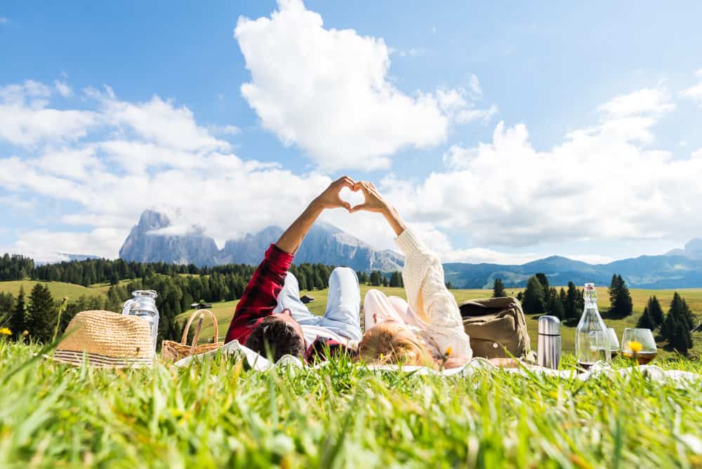 Couple laying on their backs in a field under a blue, cloudy sky while making a heart with their hands.