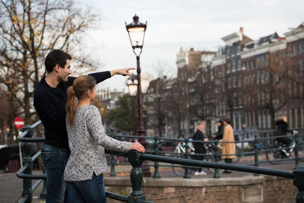 A couple is sightseeing on their European babymoon in Amsterdam with large buildings and trees in the background