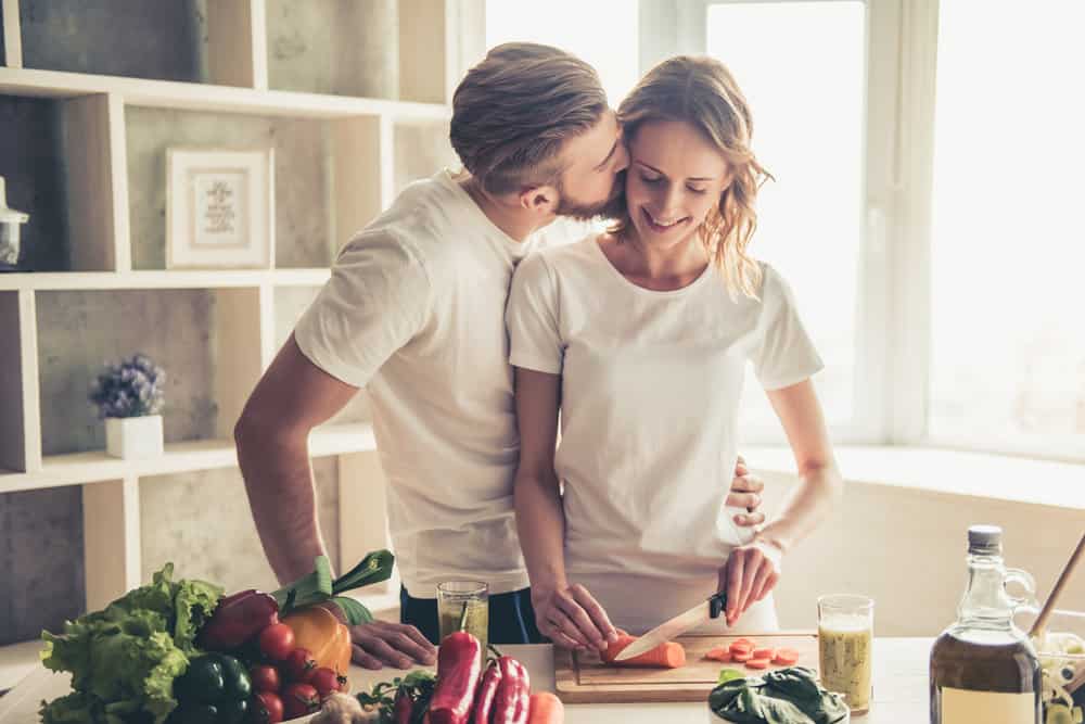 A man kisses his spouse on the cheek as she chops vegetables in a sunny kitchen while discussing how to manage expectations in marriage