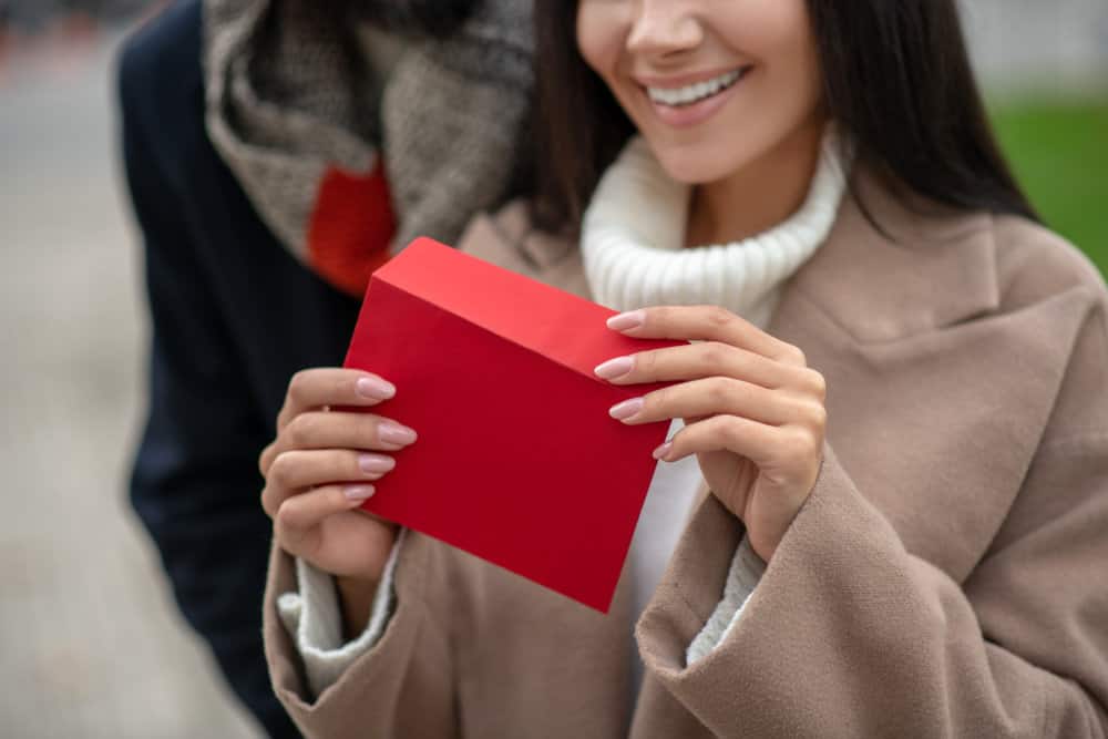 A close up of a woman smiling as she clutches a red envelope with a love letter in it as a person stands closely behind her