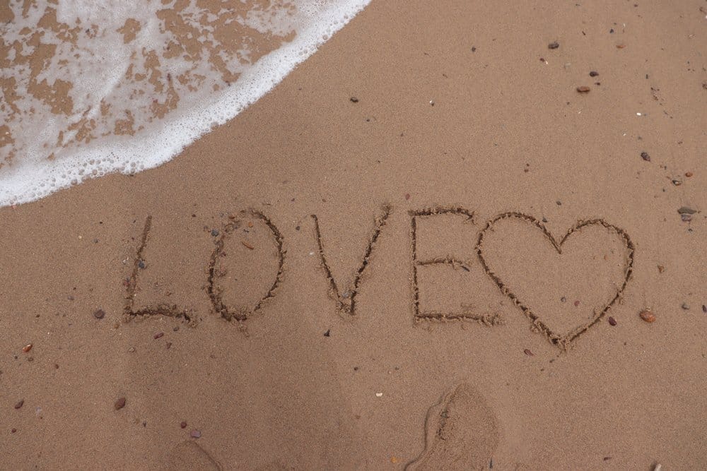 The word "Love" has been written in sand on the shore with the surf coming close to it