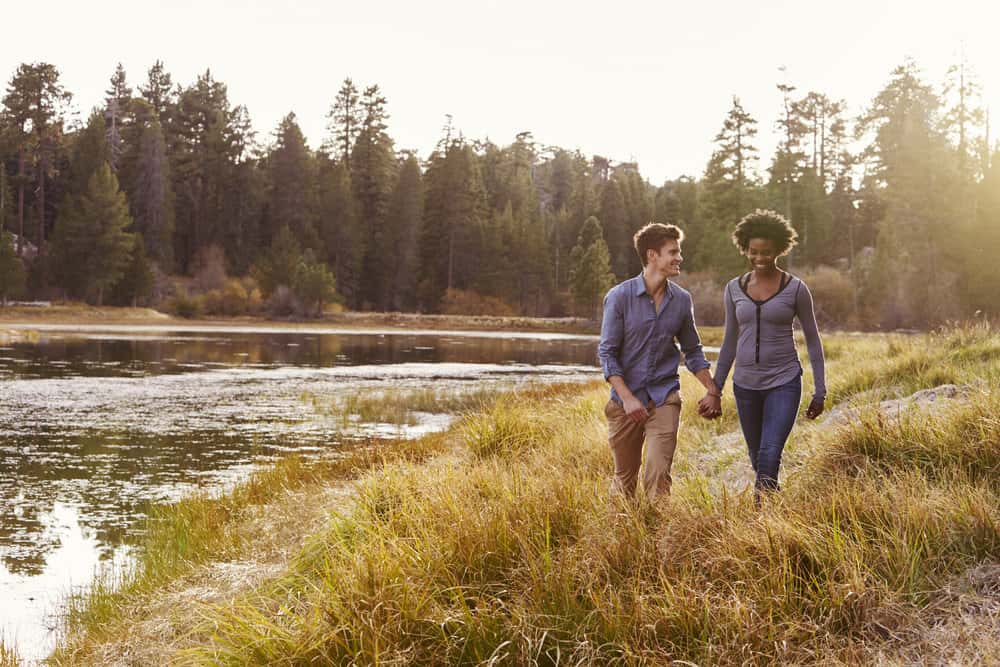 A couple holds hands as they walk beside a river with trees on the shore