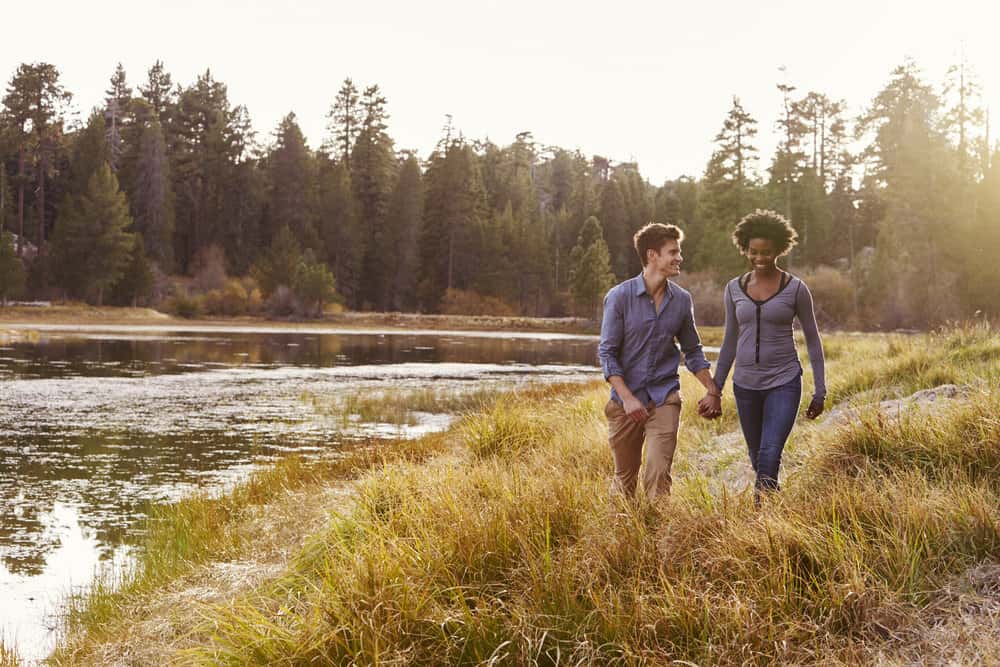 A couple holds hands as they walk abreast a river with trees on the shore