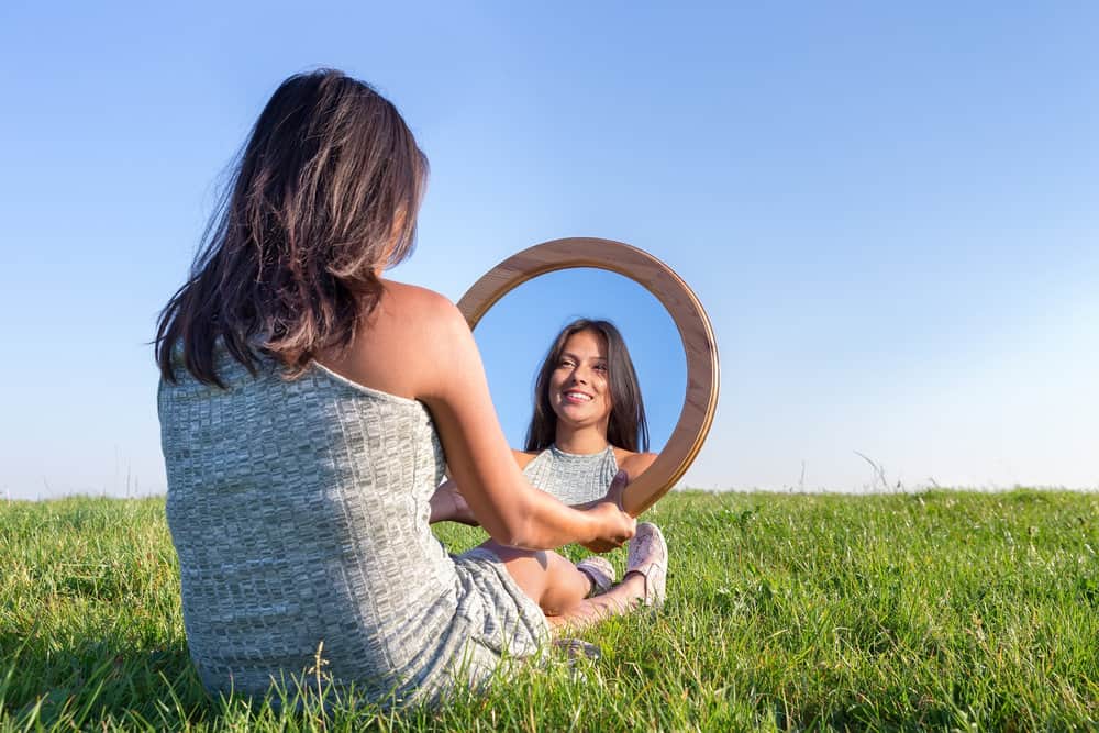 A woman sits in the grass, looking at her mirror image as she recites positive relationship affirmations