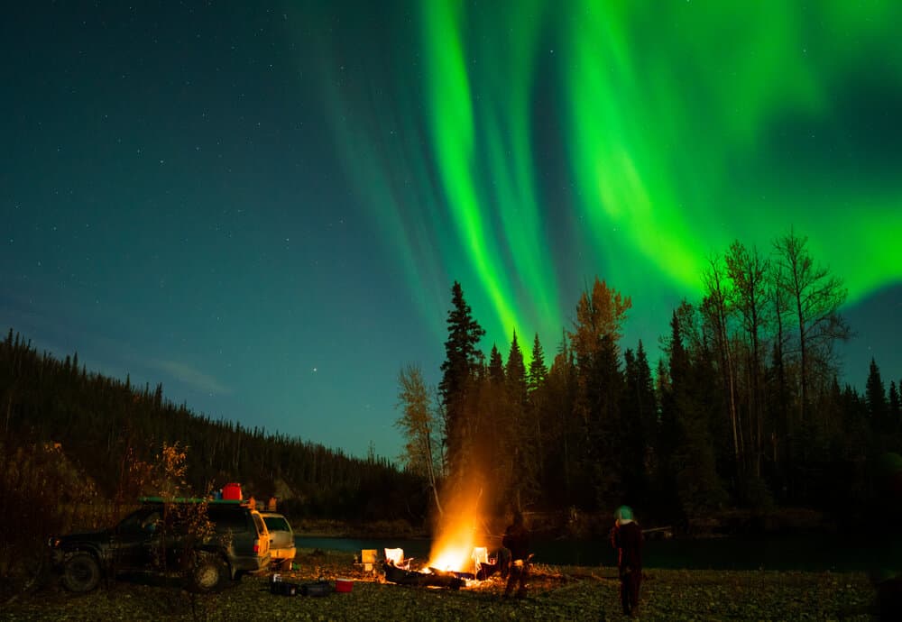The aurora borealis with neon green is seen beyond a treeline where people are camping