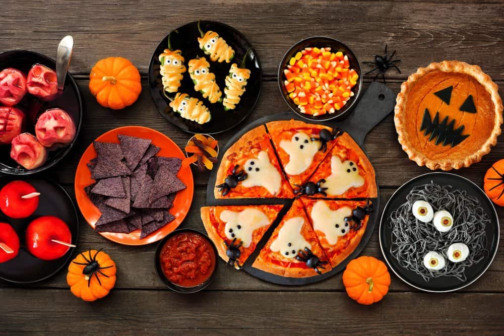 Fun Halloween dinner party table scene over a dark wood background. Top view. Pizza, jack o lantern pumpkin pie, candy apples, eyeball spaghetti, snacks and spooky punch.